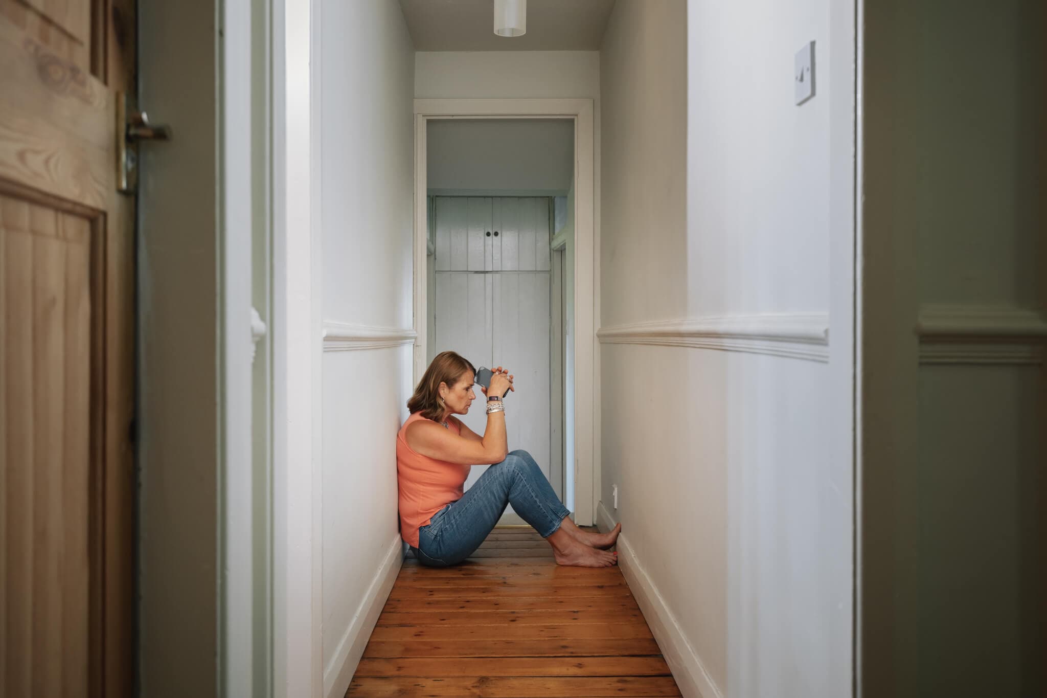 A woman in an orange tank top and jeans sits on the wooden floor of a narrow hallway, hugging her knees. - Home Instead