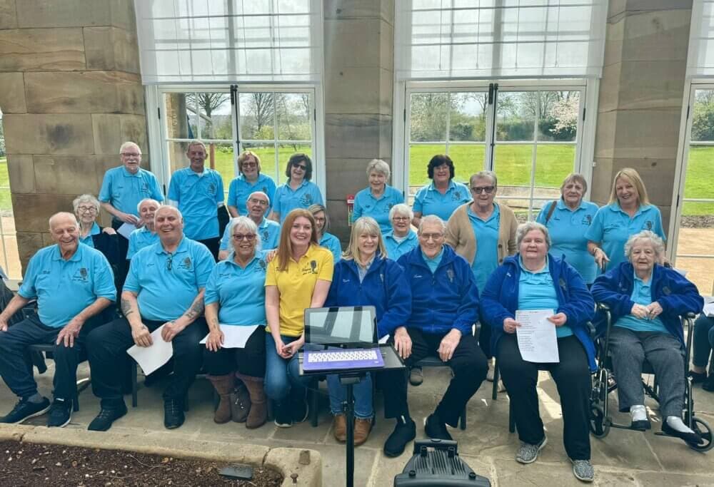 A group of elderly individuals in blue shirts posing indoors with a caregiver in a yellow shirt. - Home Instead