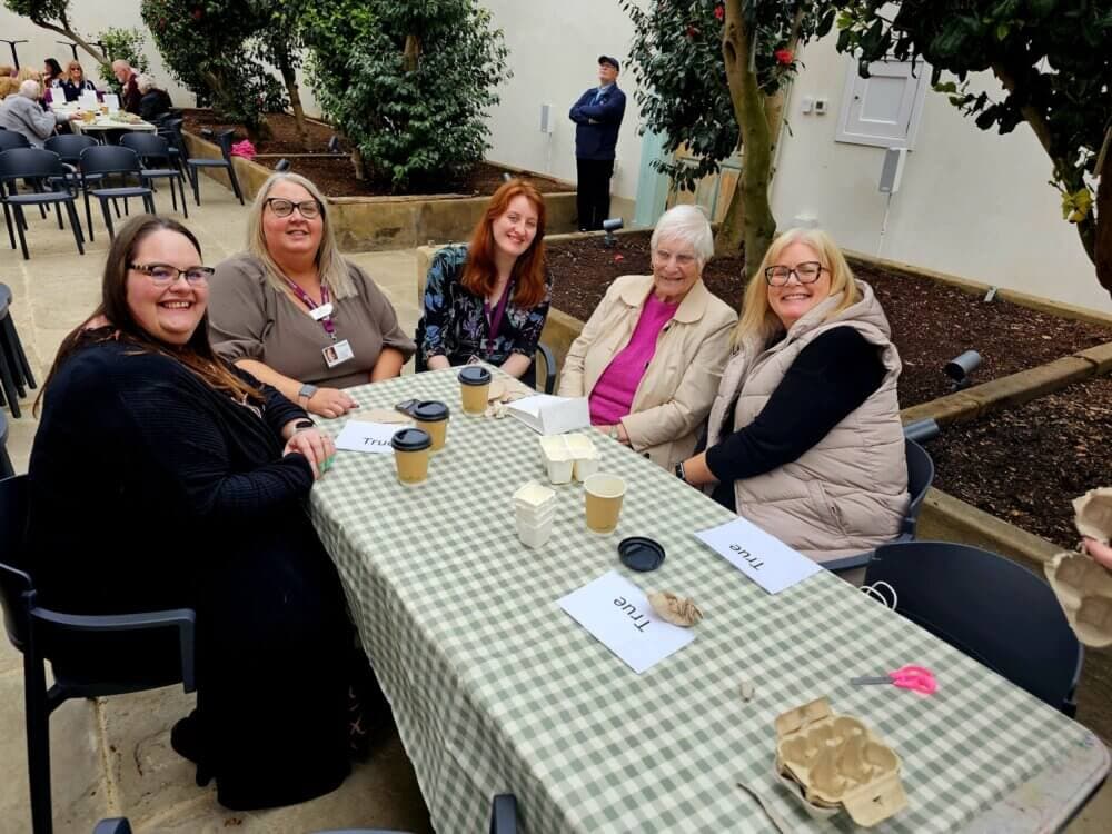 Five women sitting around a table with a green checkered tablecloth, smiling at the camera in an outdoor setting. - Home Instead
