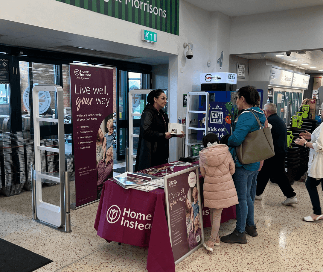 A woman at a Home Instead booth in a store talks to another woman with a child, promoting elder care services. - Home Instead