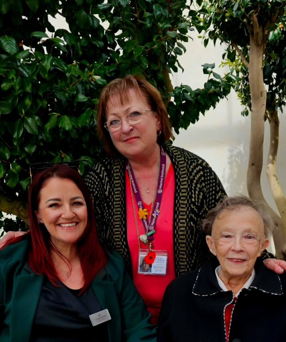 Three women are posing together, with the woman in the middle standing and the other two seated. There are trees behind them. - Home Instead