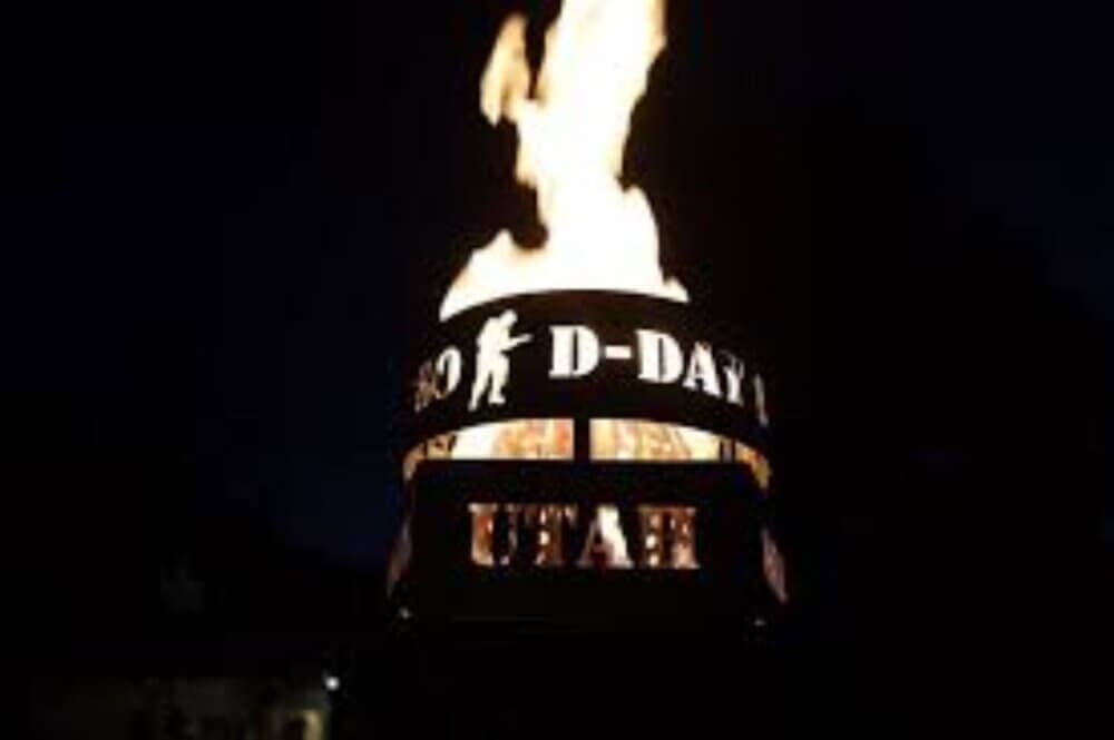 A bonfire burns inside a metal structure with cut-out letters reading "D-Day" and "Utah" against a dark background. - Home Instead
