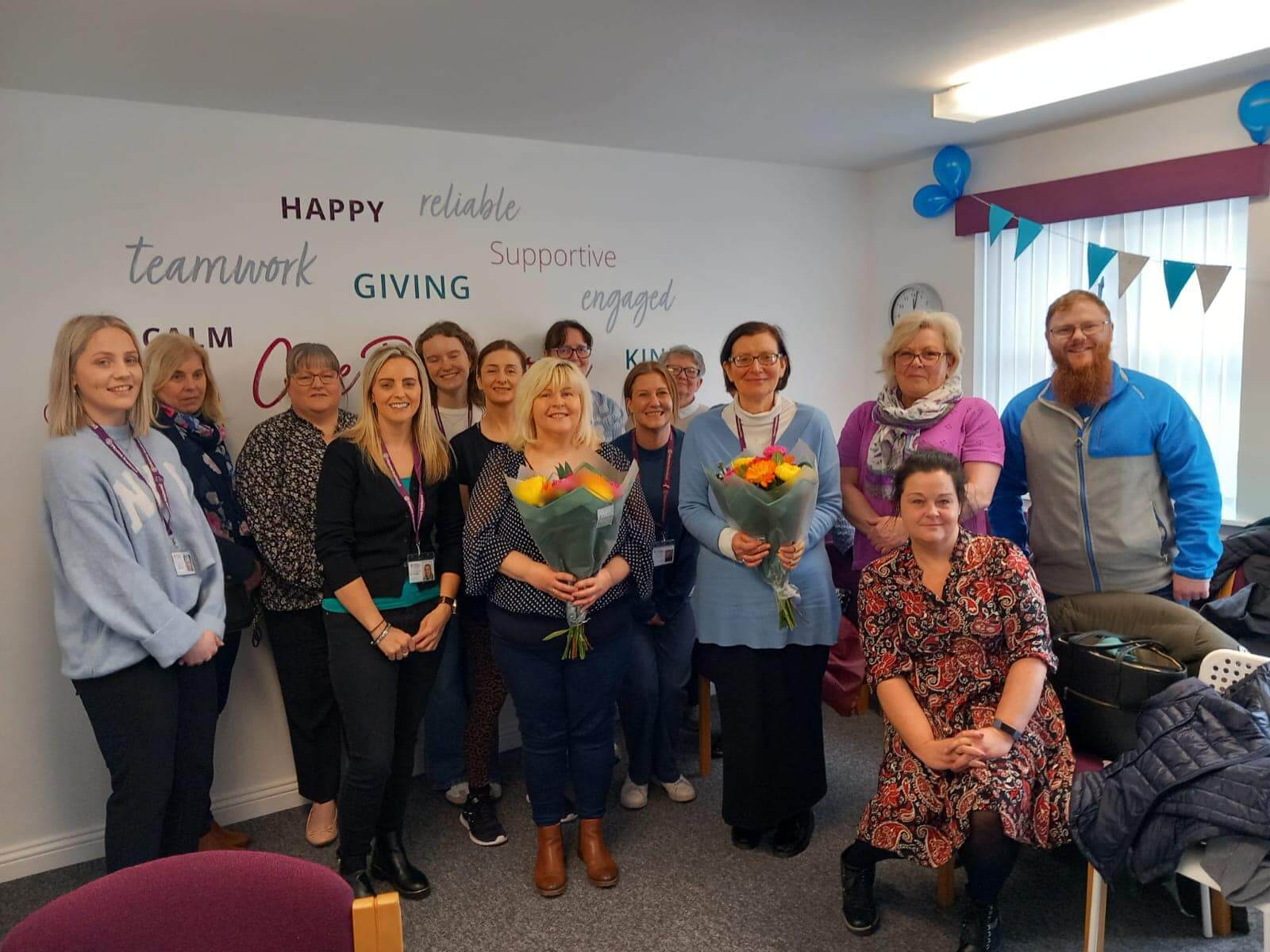 A group of people smiling indoors, two holding bouquets, with positive words on wall behind them. - Home Instead