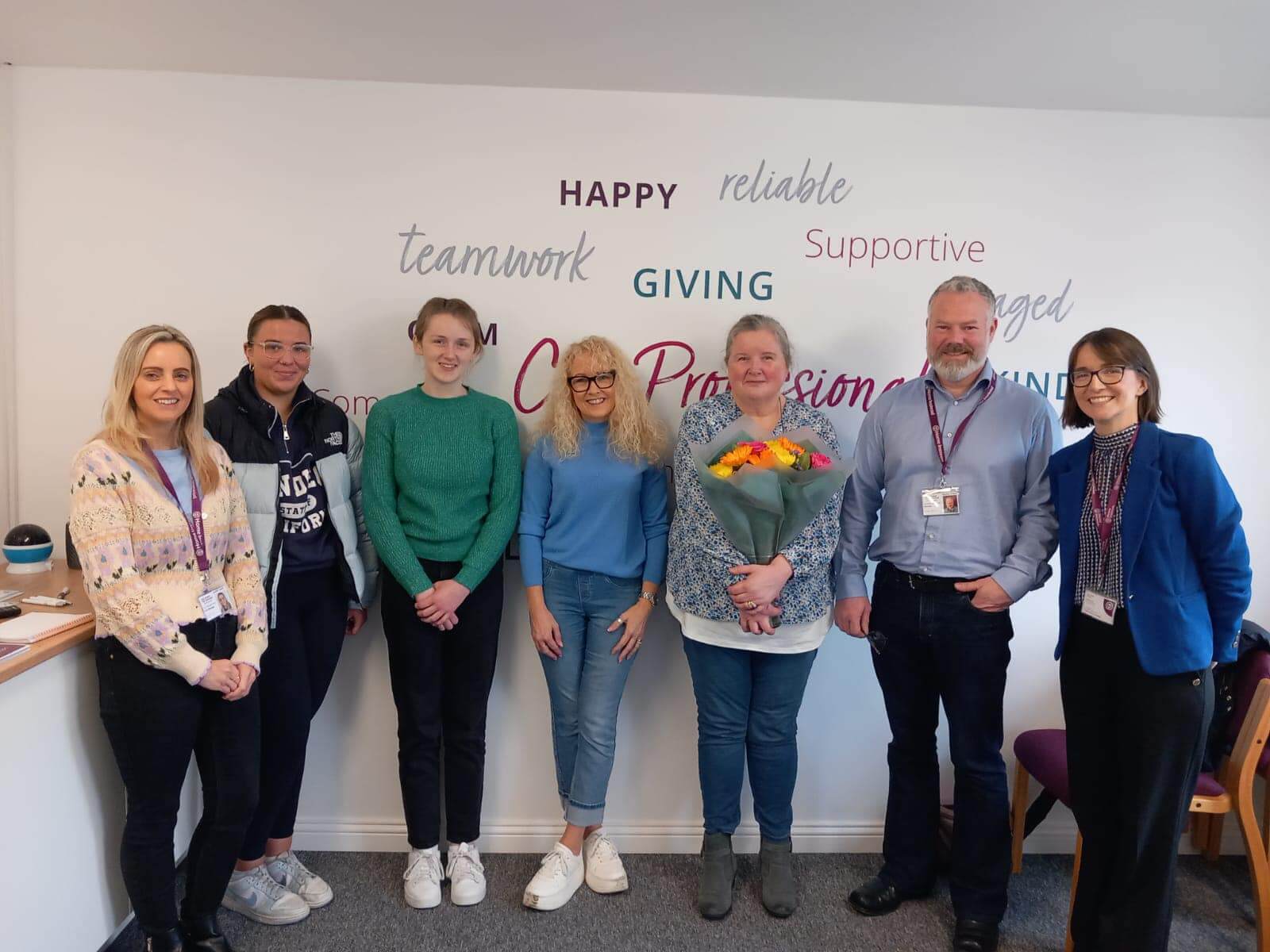 A group of seven people standing in front of a wall with motivational words. One person holds a bouquet of flowers. - Home Instead