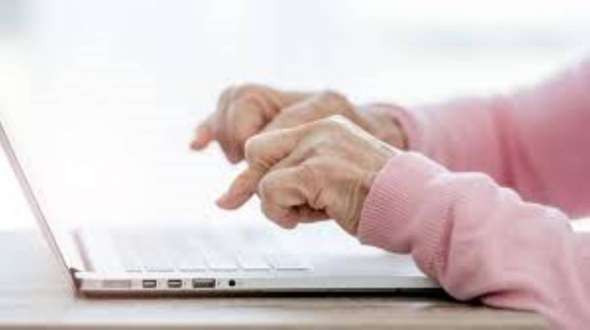 Elderly person's hands typing on a laptop keyboard, wearing a light pink sweater, with a light background. - Home Instead