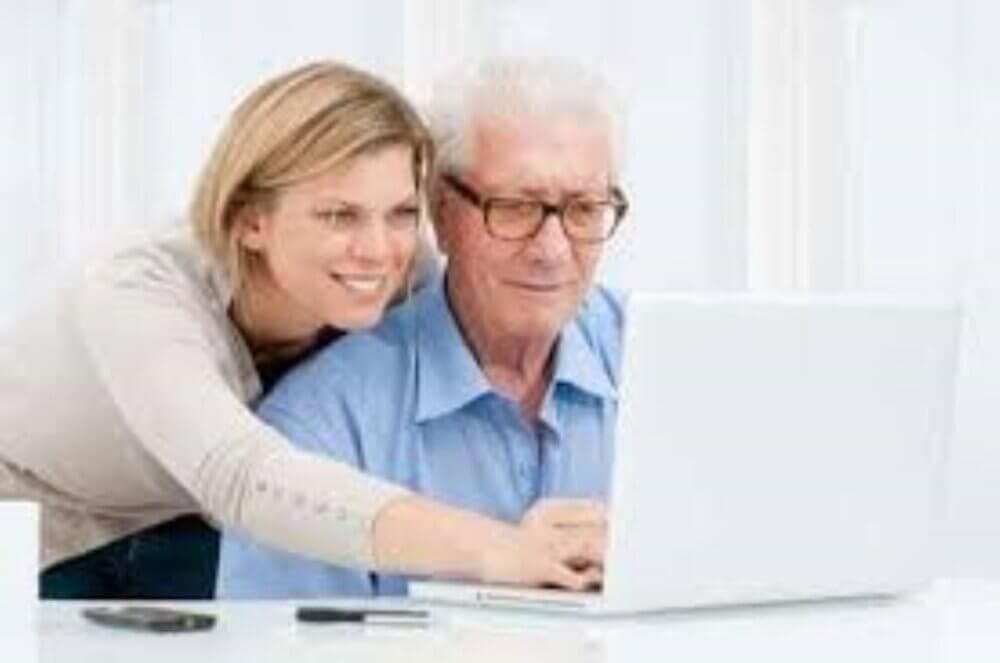 A smiling woman helps an elderly man use a laptop, both looking at the screen, with a phone and pen on the table. - Home Instead