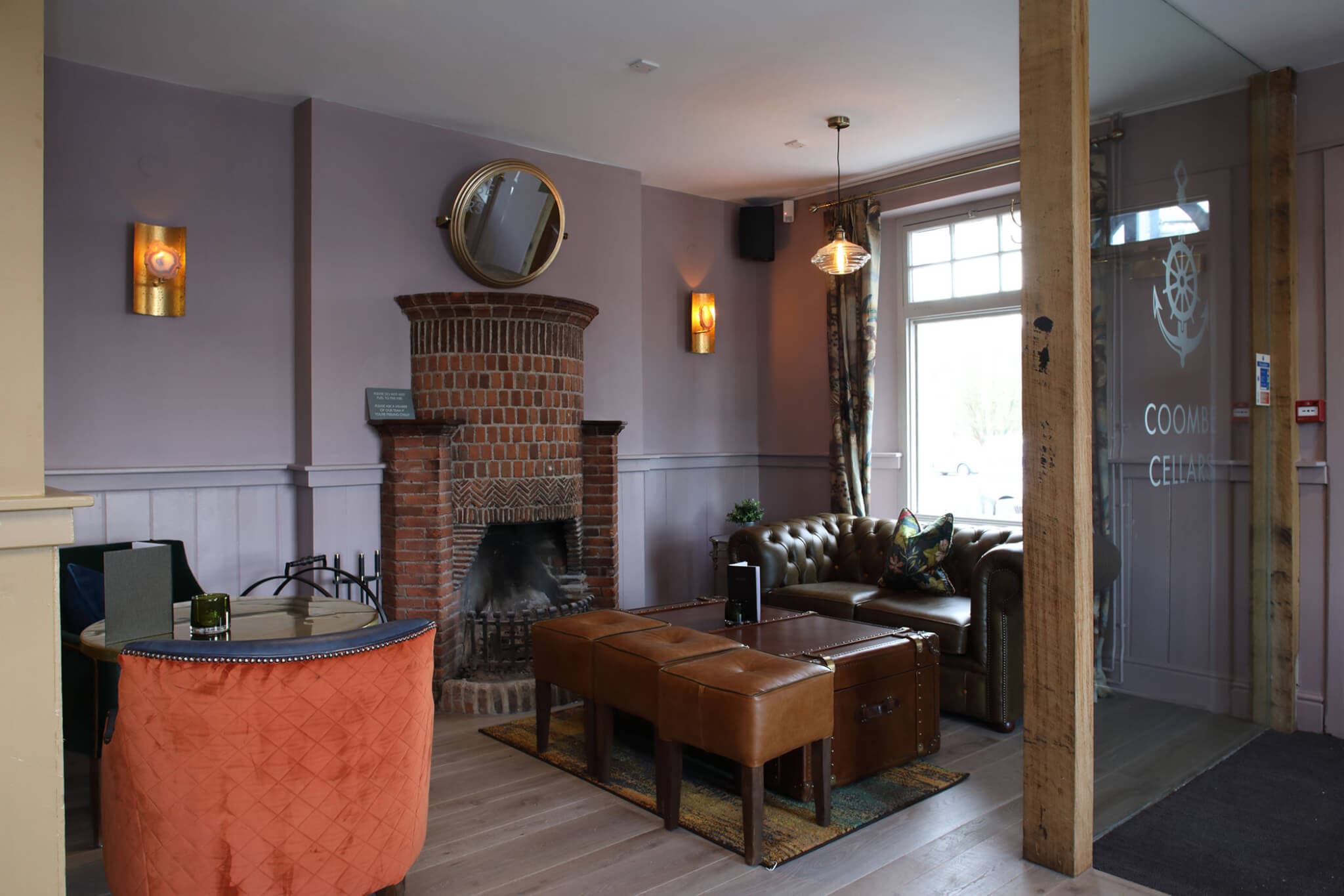 Cozy lounge area with a fireplace, leather seating, wooden stools, round mirror, and wall lights in a modern setting. - Home Instead