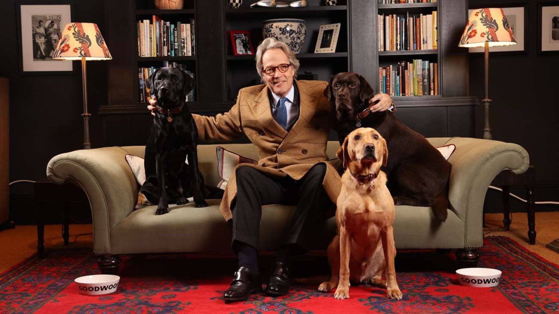 An older man sits on a couch with three dogs, two black and one yellow, in a cozy room with bookshelves. - Home Instead