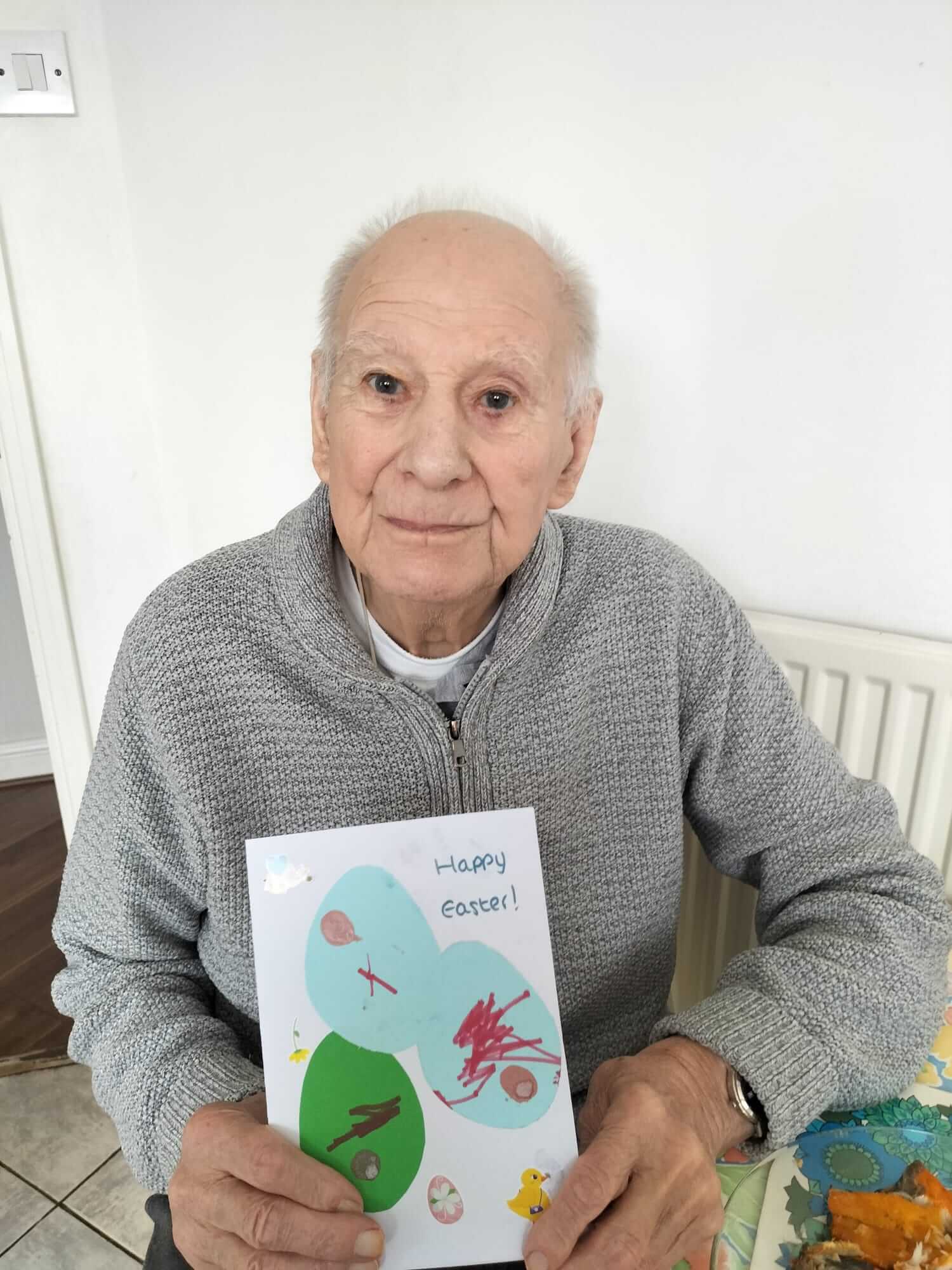 Elderly man smiling and holding a handmade Easter card with colorful decorations and "Happy Easter!" text. - Home Instead