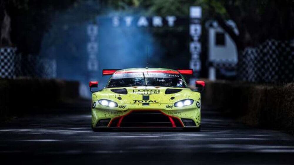 A lime-green race car speeds down a track with a "START" sign overhead, amid surrounding trees and a smoky background. - Home Instead