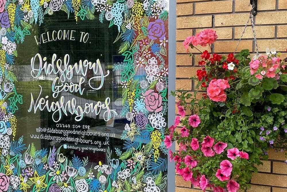 Sign with "Welcome to Didsbury Good Neighbours" surrounded by floral drawings, next to a hanging basket of pink flowers. - Home Instead