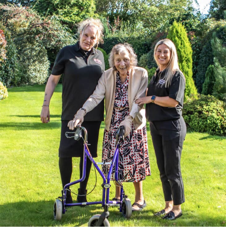 Two caregivers smiling, assisting an elderly woman with a walker on a sunny garden lawn. - Home Instead