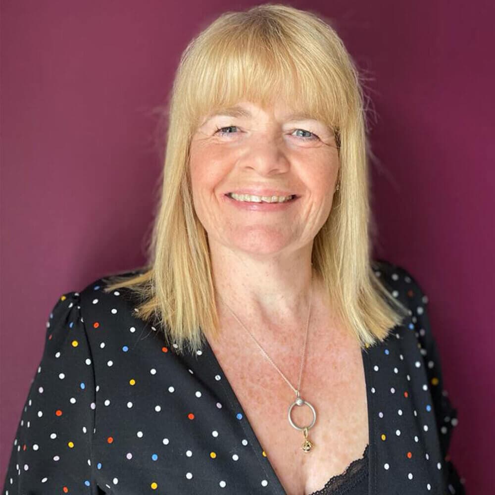 Smiling woman with blonde hair wearing a polka dot dress and a necklace, standing against a purple background. - Home Instead