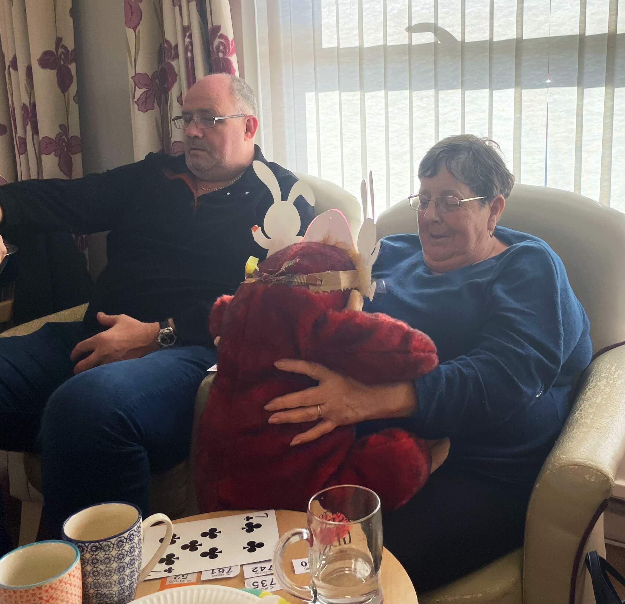 Two seated adults holding a teddy bear with reindeer antlers. Nearby are a deck of cards and coffee mugs on a table. - Home Instead