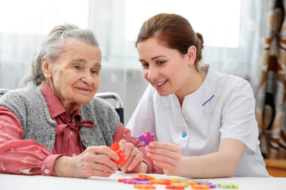 An elderly woman and a caregiver smile and work together on a puzzle at a table indoors. - Home Instead