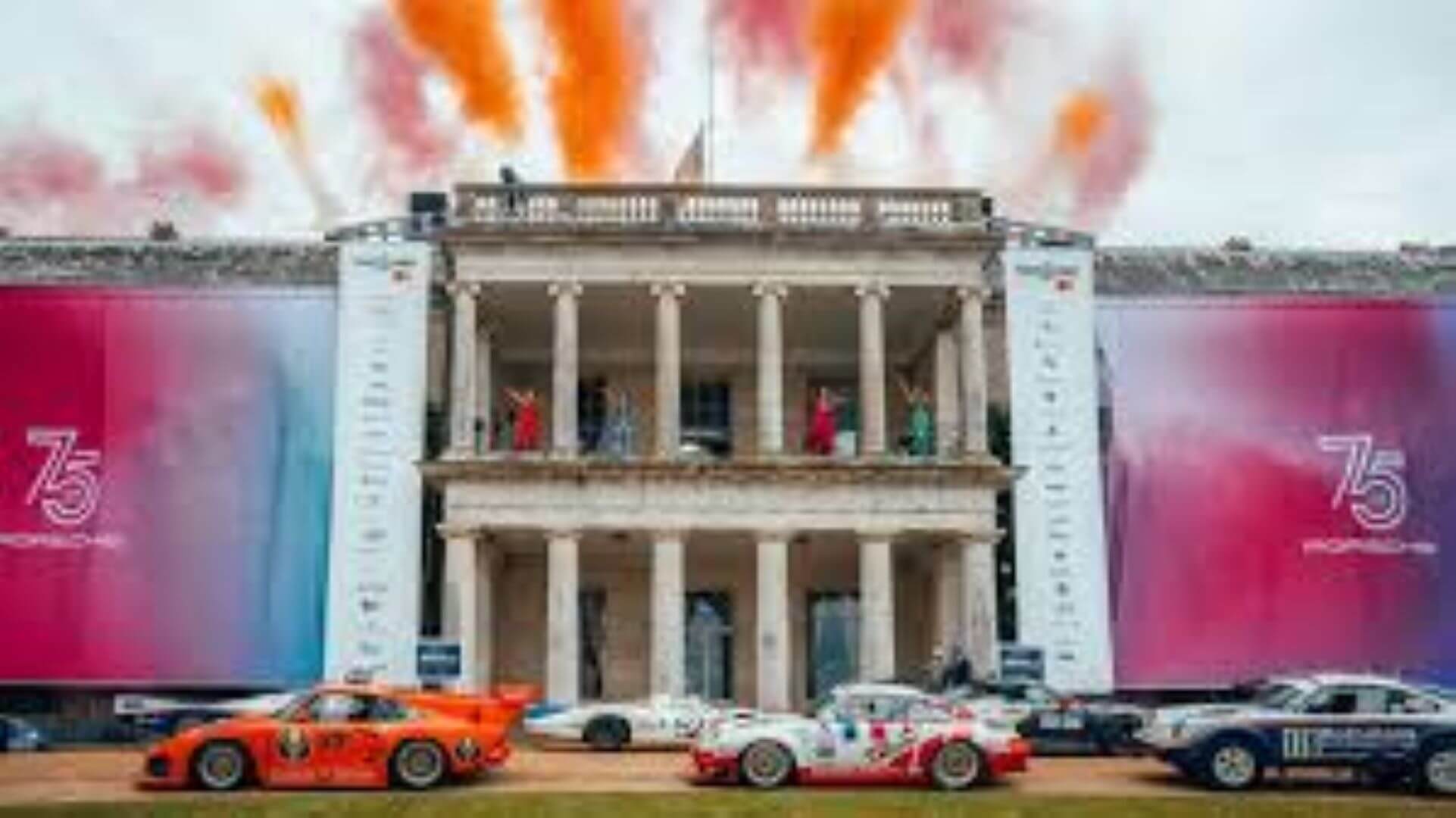 Porsche 75th anniversary celebration with cars in front of a grand building and vibrant orange fireworks overhead. - Home Instead
