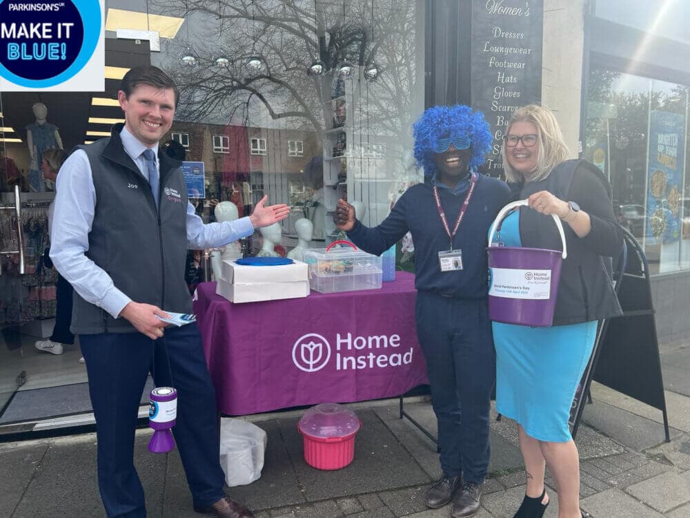 Three people fundraising, one in a blue wig, stand in front of a "Home Instead" table with donation buckets. - Home Instead
