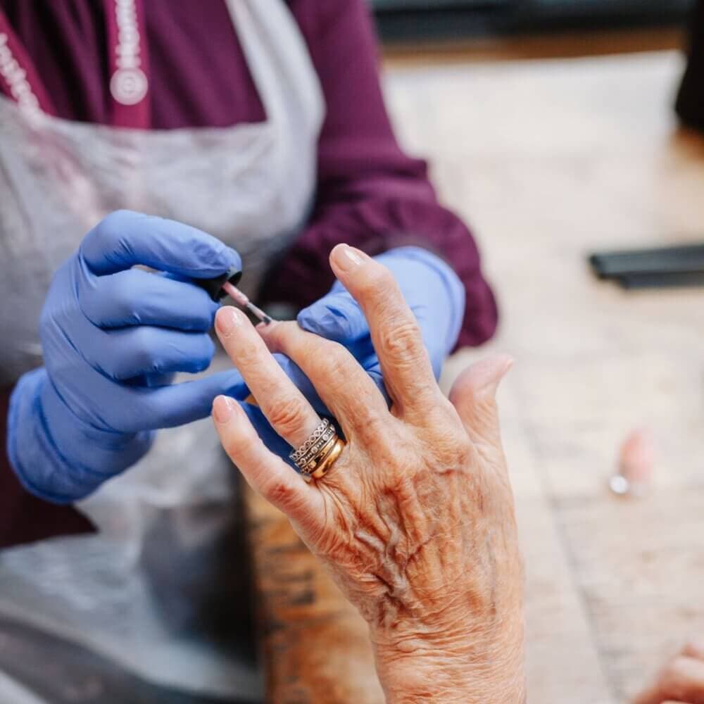 A person wearing blue gloves applies nail polish on the nails of an elderly person's hand. - Home Instead