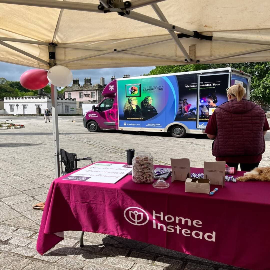 A booth with a pink Home Instead tablecloth set up outside near a parked dementia awareness tour van. - Home Instead