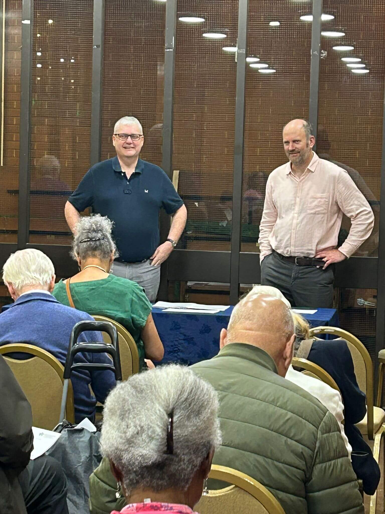 Two men stand at the front of a room addressing an audience seated in rows. - Home Instead