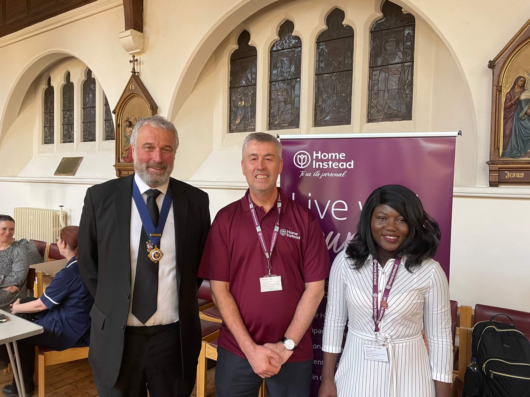 Three people posing for a photo in front of a "Home Instead" banner in a room with arched windows and religious artwork. - Home Instead
