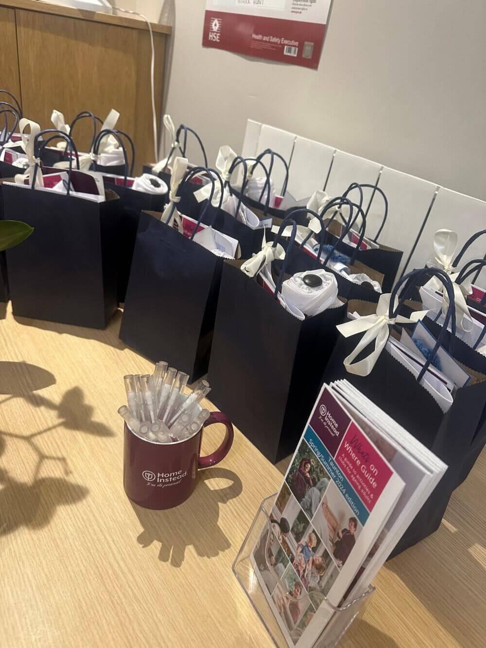 Several navy gift bags with white ribbons, a branded mug with pens, and a stack of brochures on a wooden table. - Home Instead