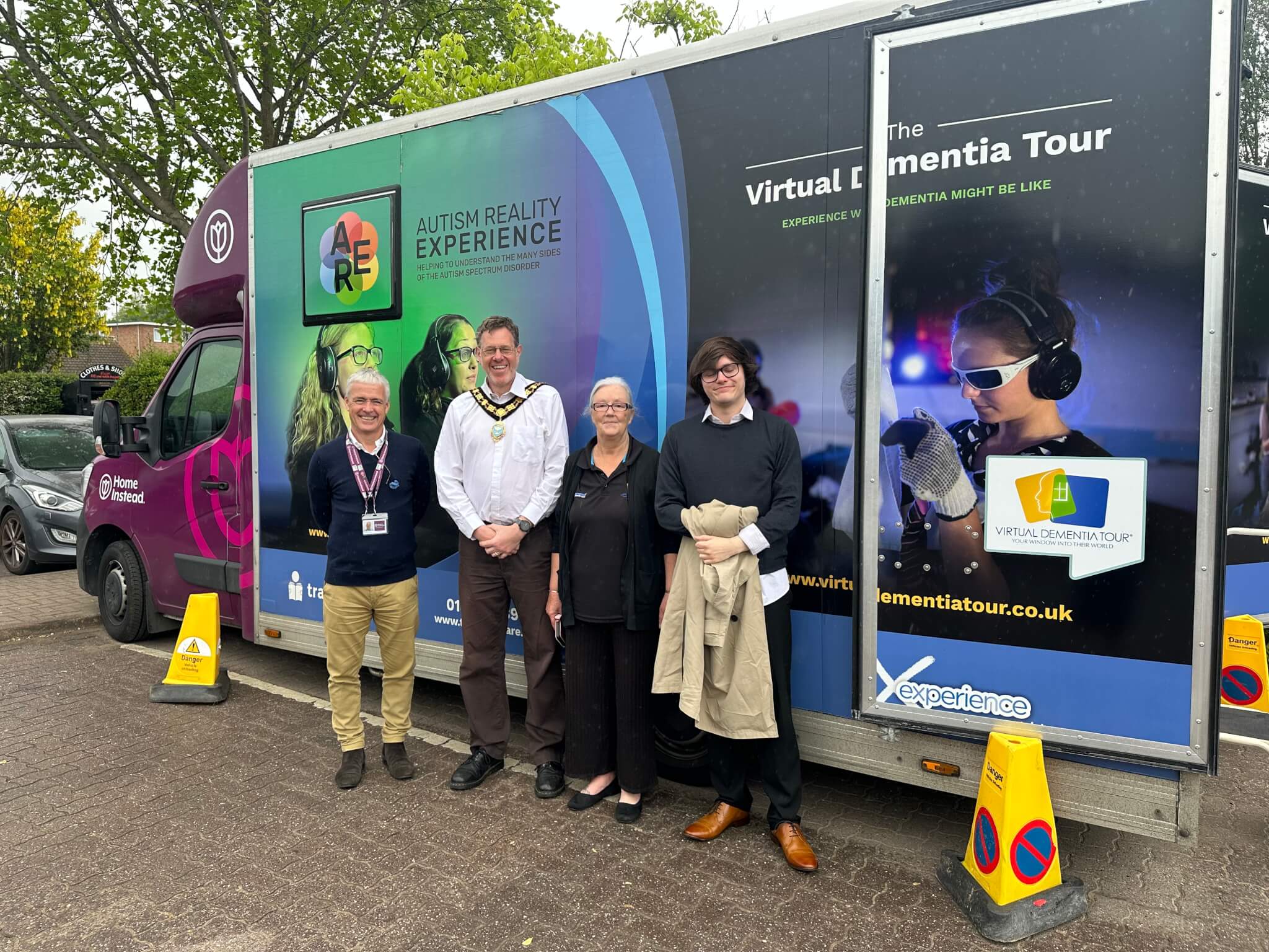Four people standing in front of a colorful Autism Reality Experience tour truck with trees in the background. - Home Instead