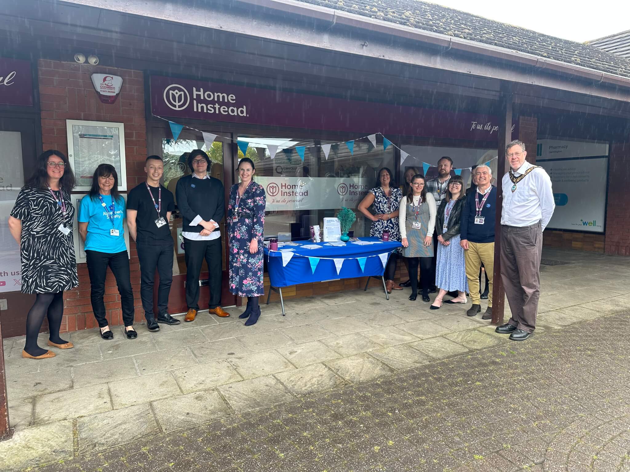 A group of people stand in front of an outdoor booth with a "Home Instead" banner and promotional materials. - Home Instead