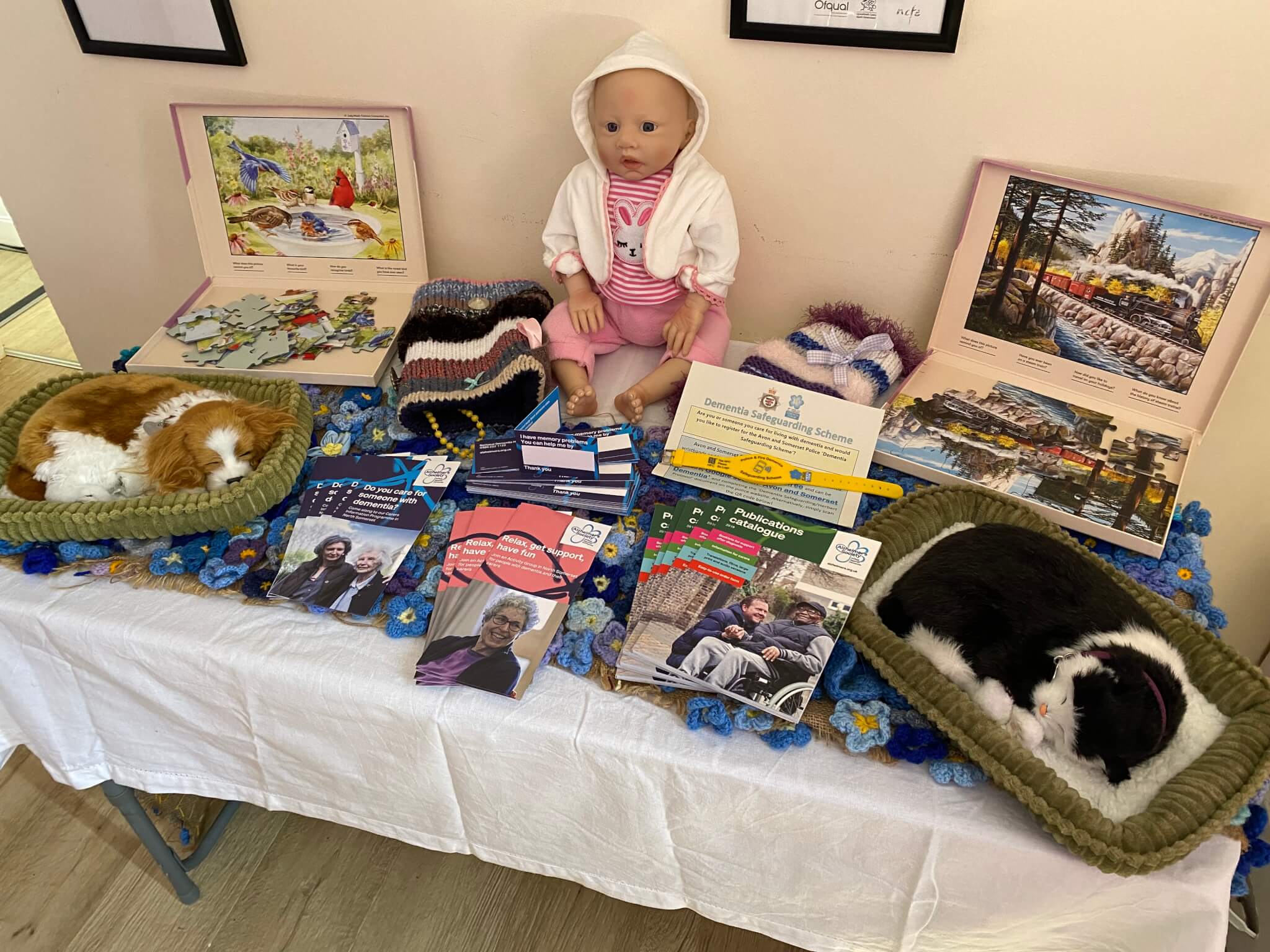 A display table with knitted hats, brochures, a doll, a jigsaw puzzle, and sleeping cats in baskets. - Home Instead