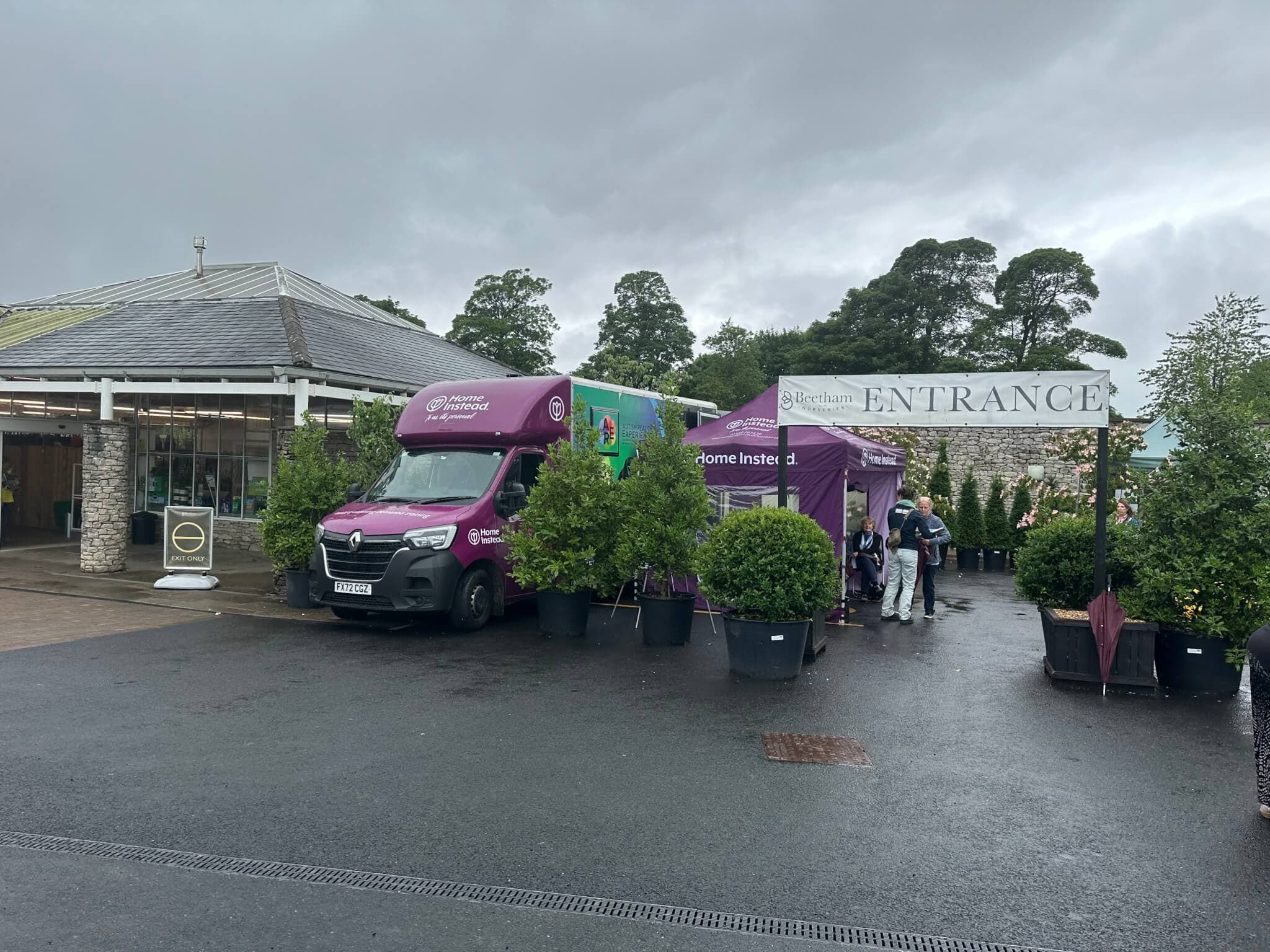 Purple van and tent near entrance of a garden center, surrounded by plants and trees, under an overcast sky. - Home Instead