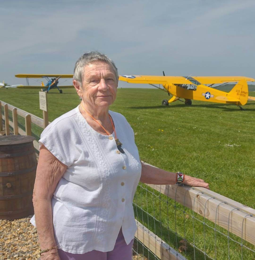 An older woman stands by a fence with yellow vintage aircraft on a grassy field in the background. - Home Instead