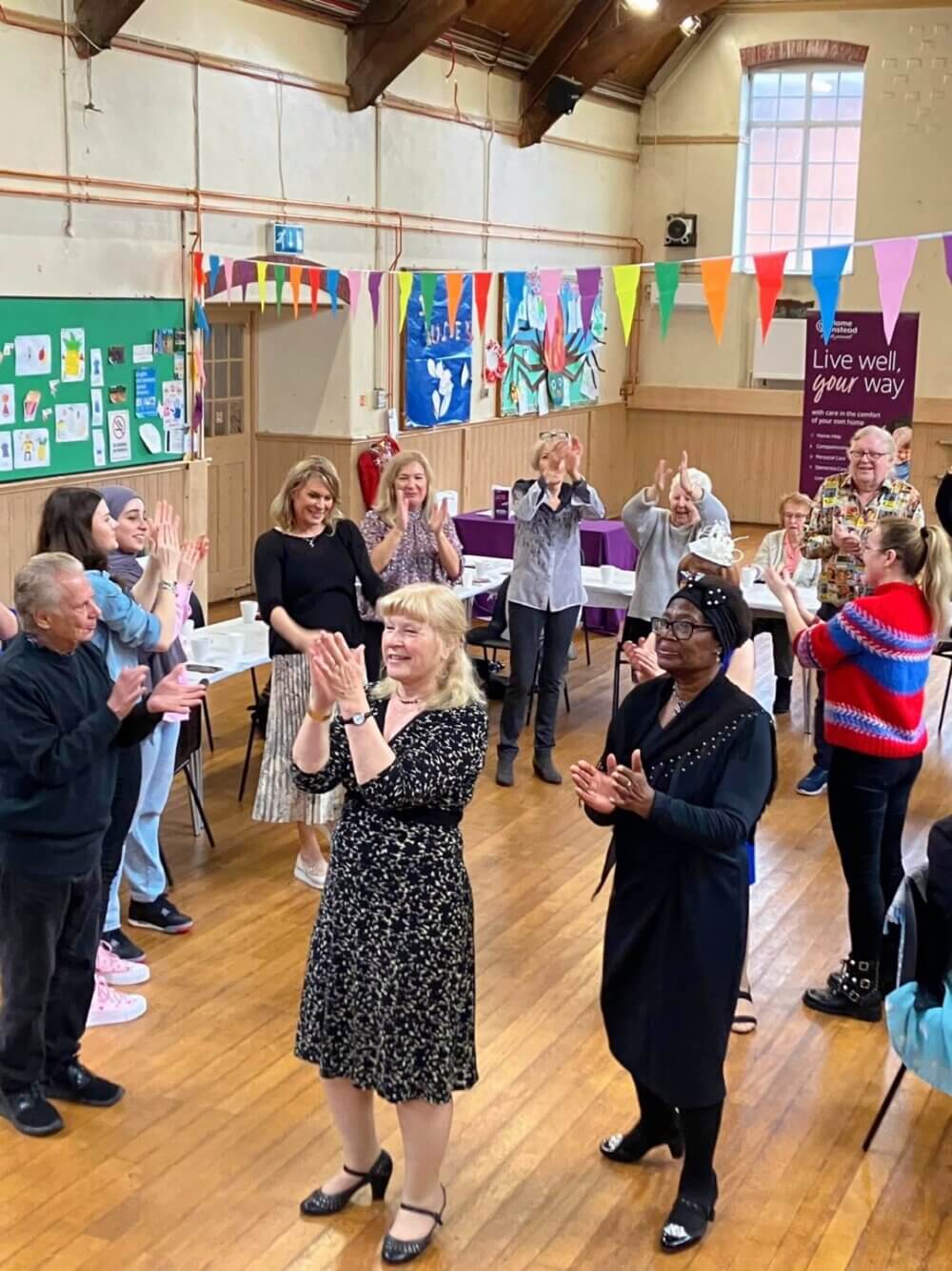 A group of people in a community hall clapping and dancing, with colorful bunting and artwork on the walls. - Home Instead
