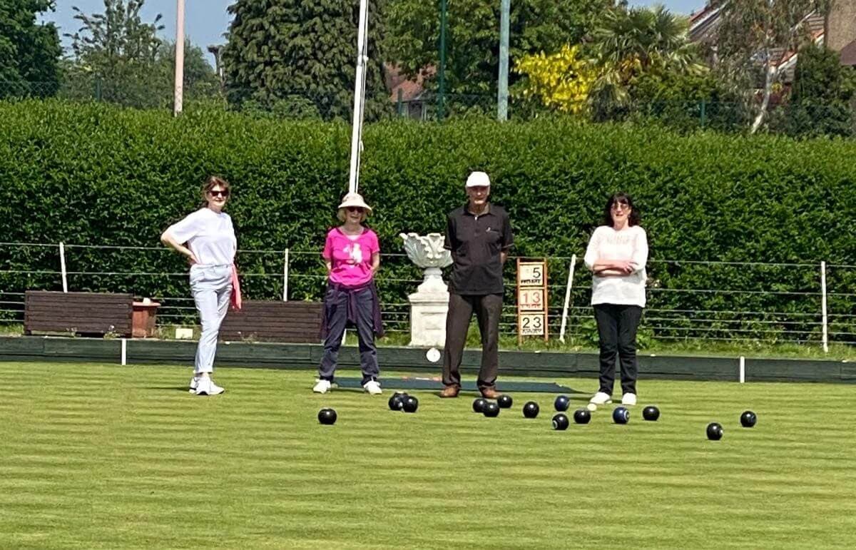 Four people playing lawn bowls on a green field, with a hedge and trees in the background on a sunny day. - Home Instead