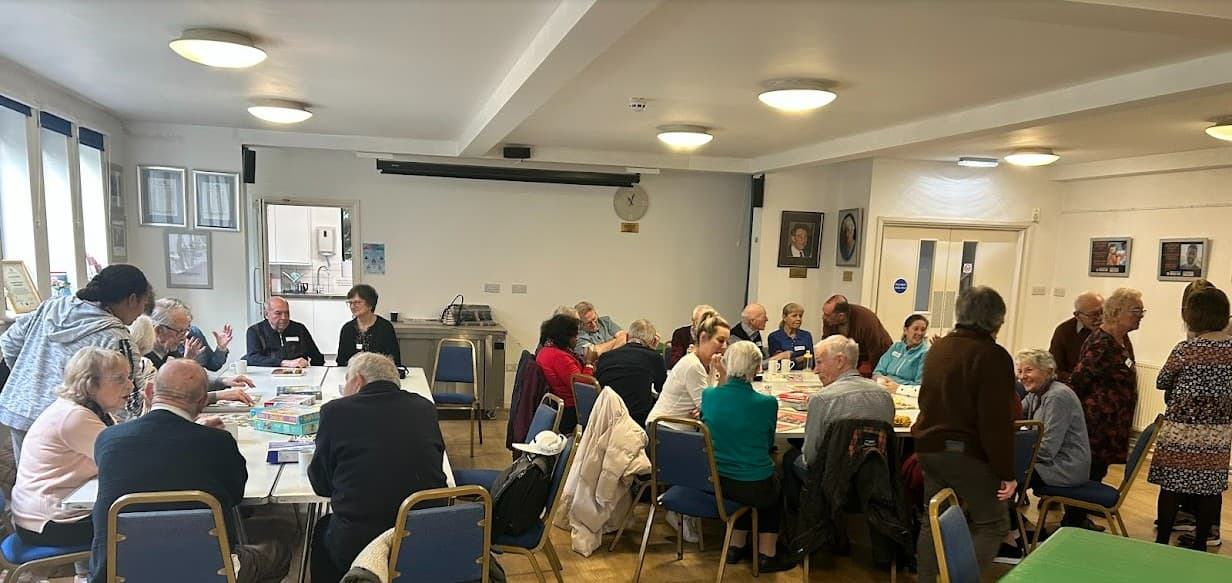 A group of older adults sit and chat around tables in a bright community room. - Home Instead