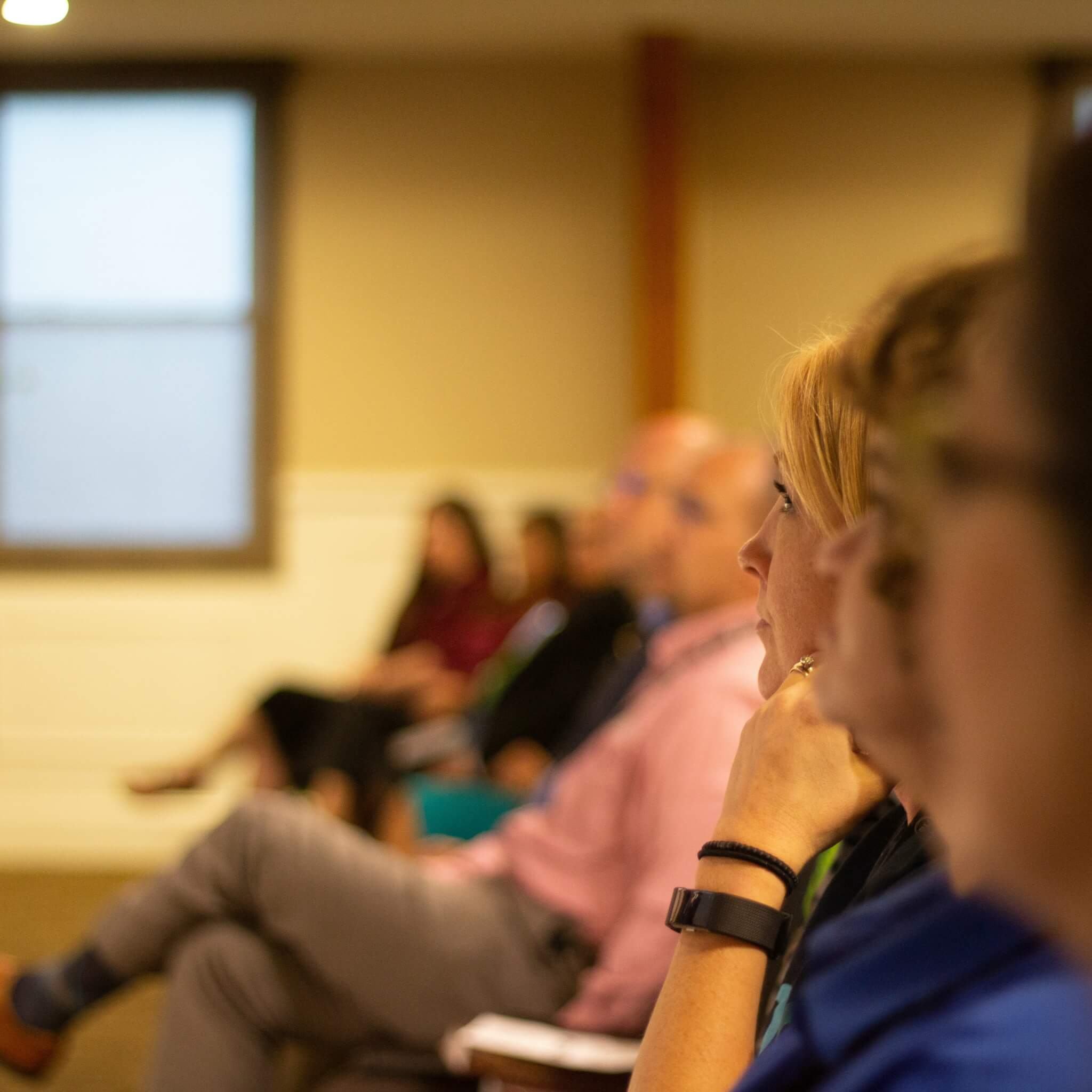 People seated and attentively listening in a dimly lit room, with a window visible in the background. - Home Instead