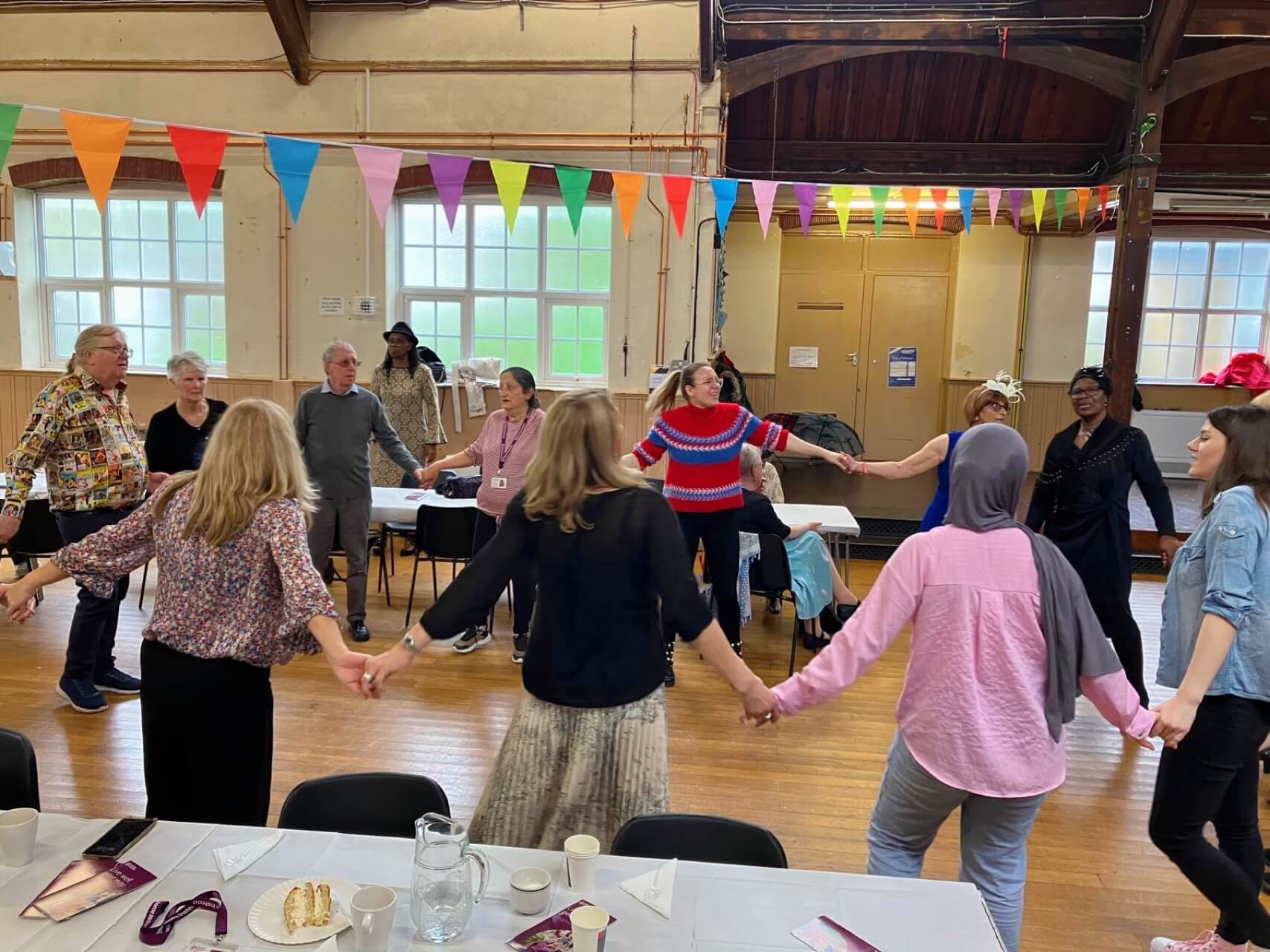 People holding hands in a circle inside a decorated hall, colorful bunting hanging overhead. Various table settings visible. - Home Instead