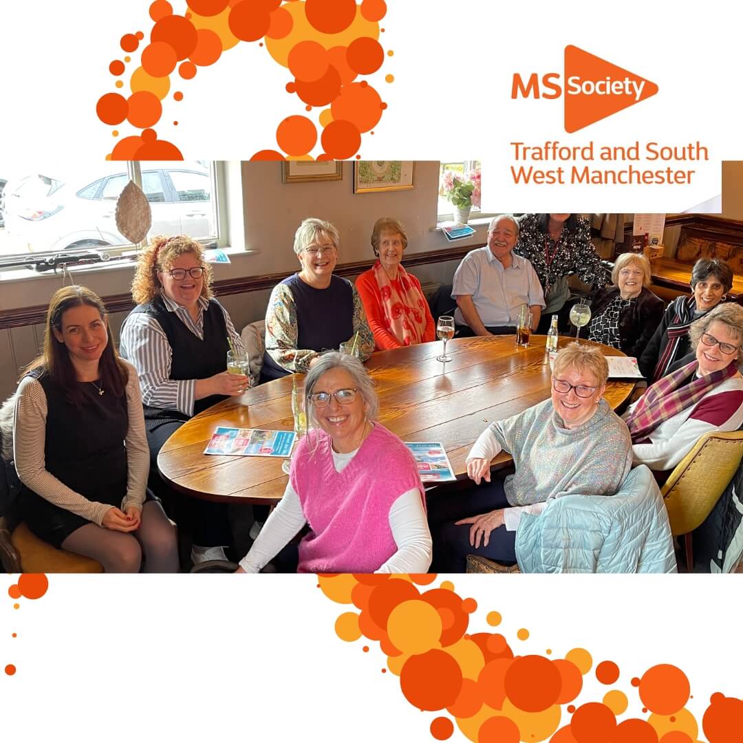A group of people sitting around a table, smiling, under an MS Society Trafford and South West Manchester banner. - Home Instead