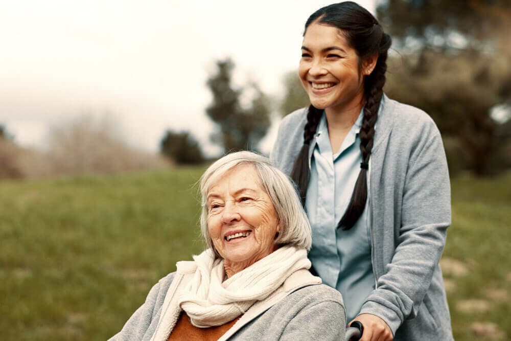 A young woman pushes an elderly woman in a wheelchair outdoors, both smiling and enjoying the day. - Home Instead