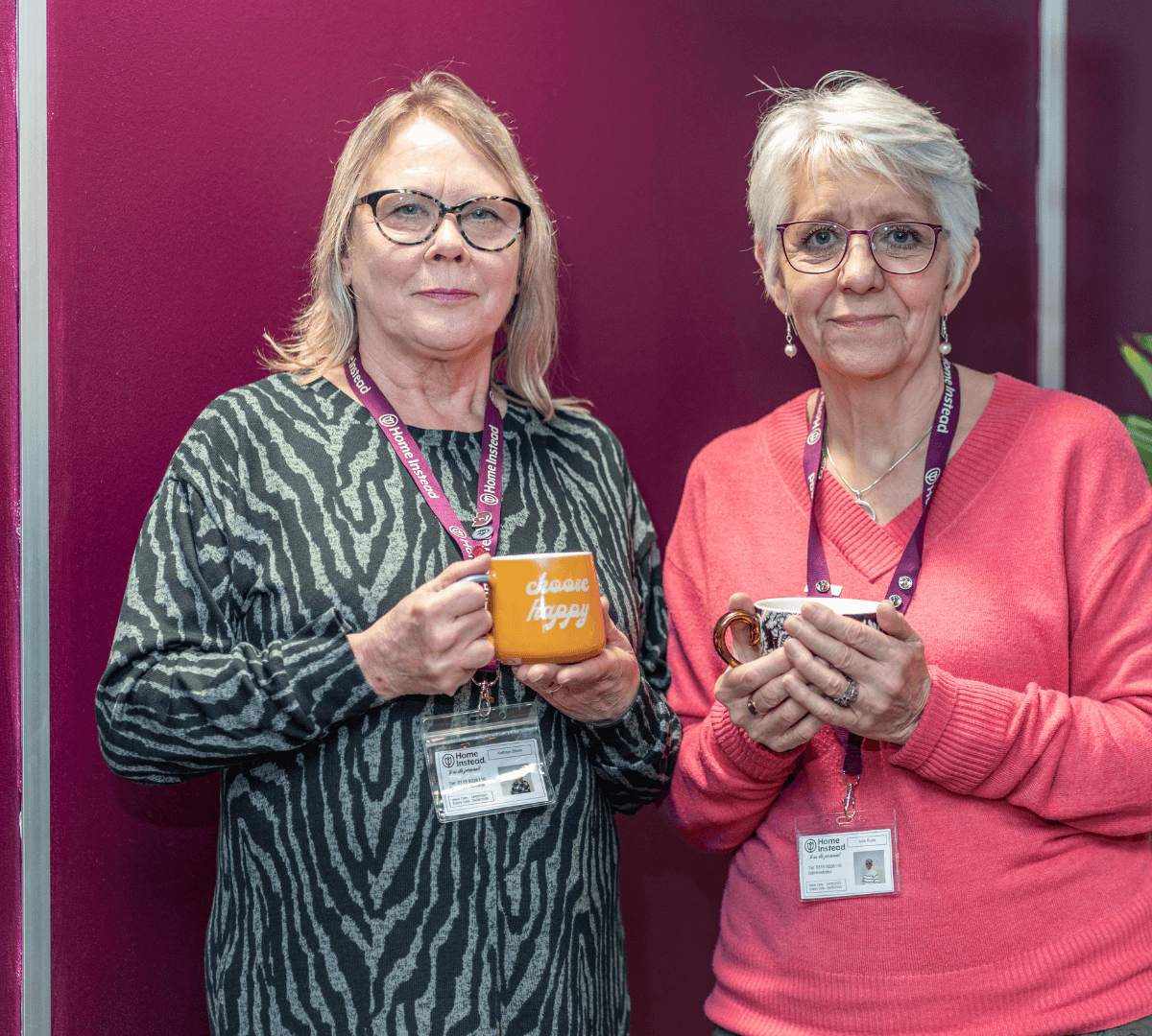 Two women who are our Care Professionals wearing our Home Instead Lanyards and both holding a cup of tea in their hands, a purple backdrop in Home Instead brand colours.