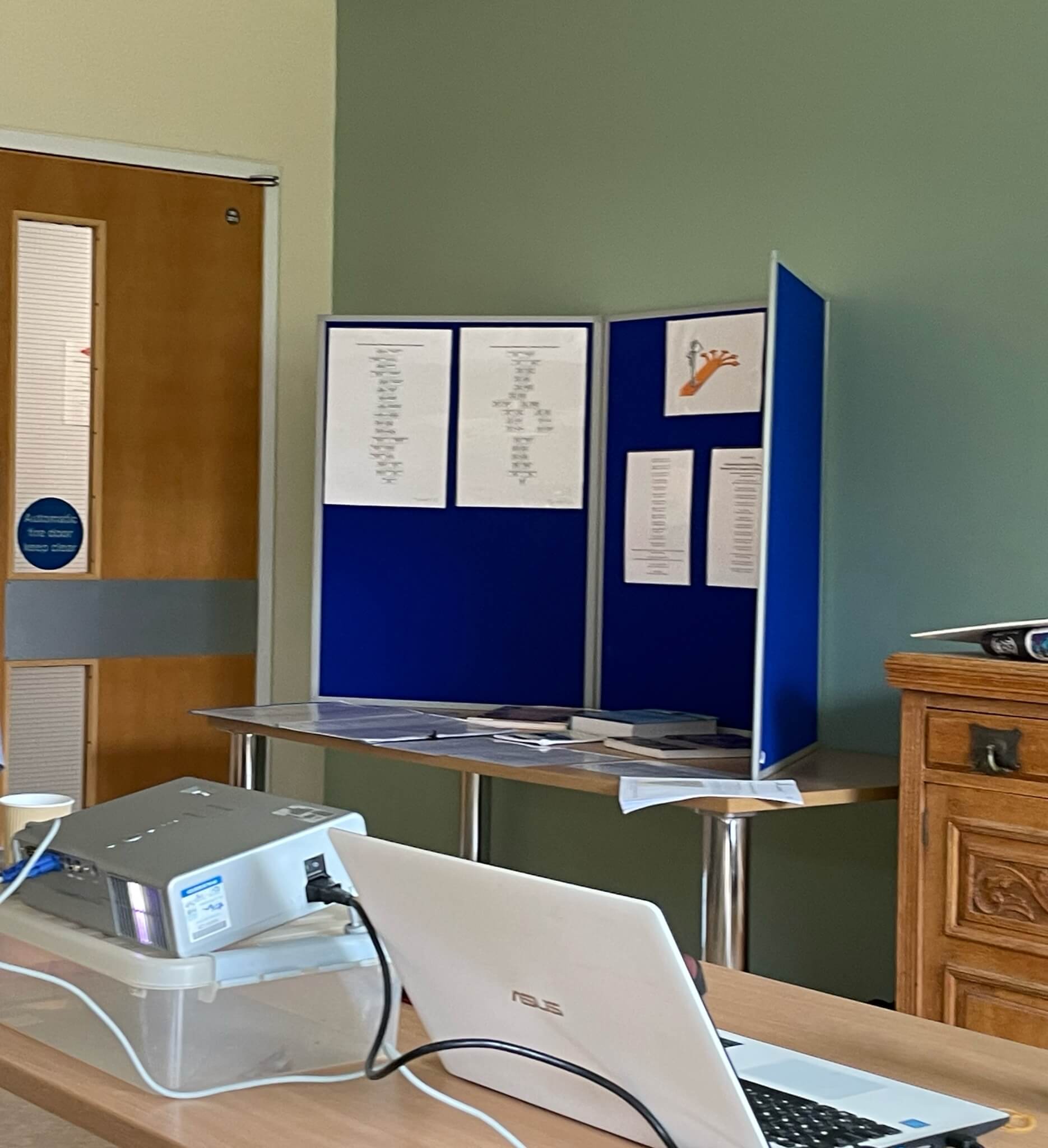 A room with a laptop, a projector, a display board with papers, and a door in the background. - Home Instead