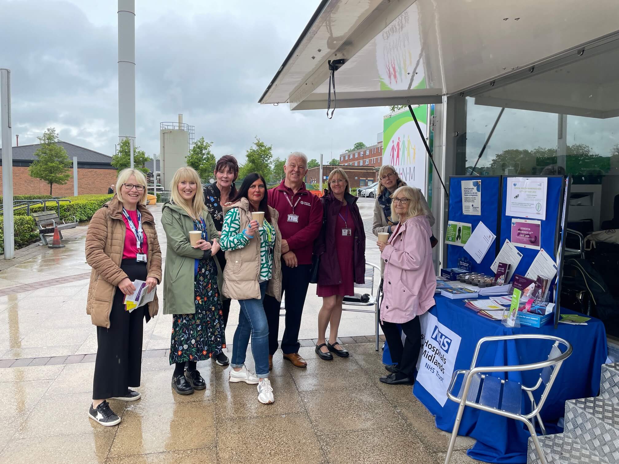 A group of people standing together outdoors beside an informational booth with pamphlets and a banner on display. - Home Instead