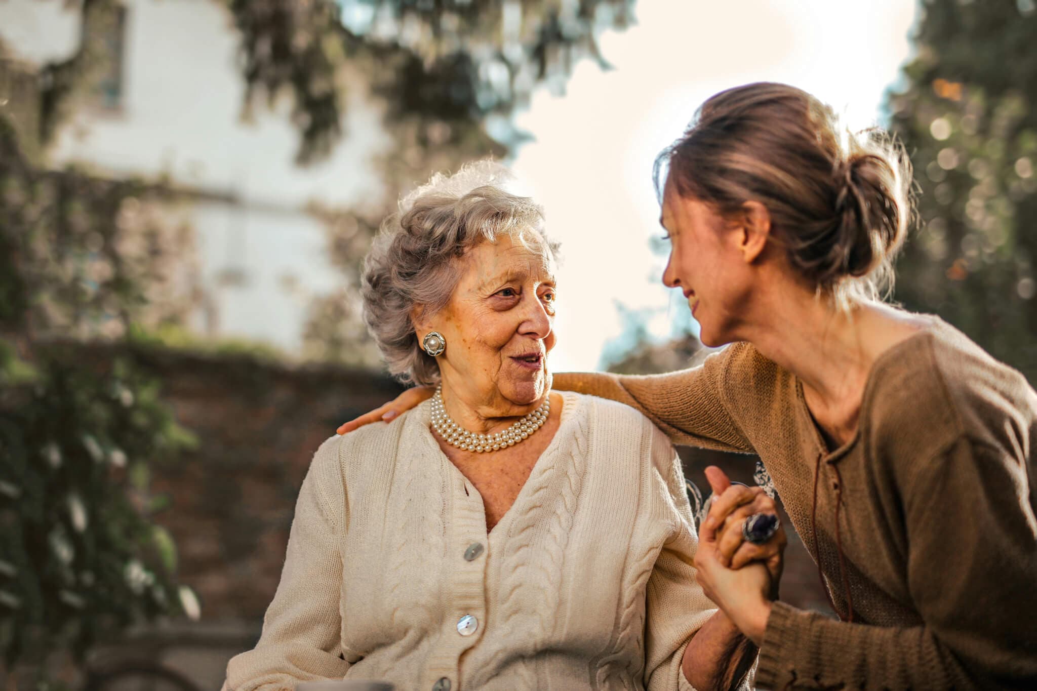 Older woman and younger woman smiling at each other warmly, sharing a moment outdoors in a garden. - Home Instead