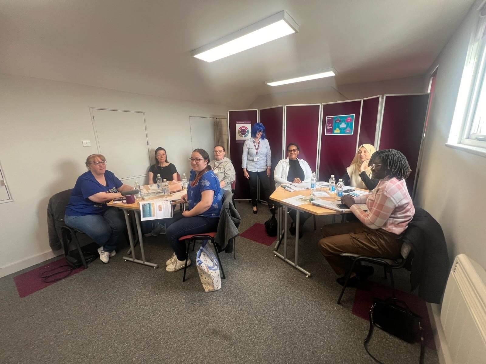 A group of people sit around tables, engaged in discussion in a room with red dividers and a whiteboard. - Home Instead
