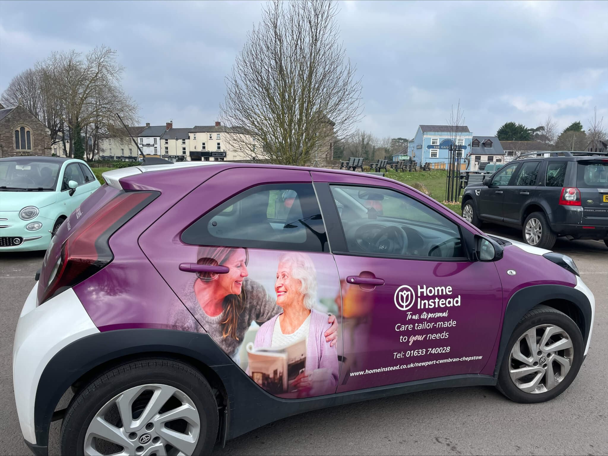 A purple and white "Home Instead" car with an elderly woman and caregiver graphic, parked in a lot on a cloudy day. - Home Instead