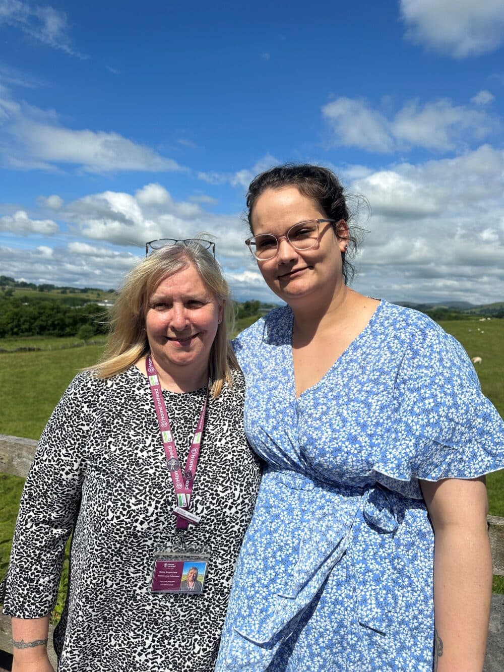 Two women stand outdoors on a sunny day, smiling; one in a black and white dress, the other in a blue floral dress. - Home Instead