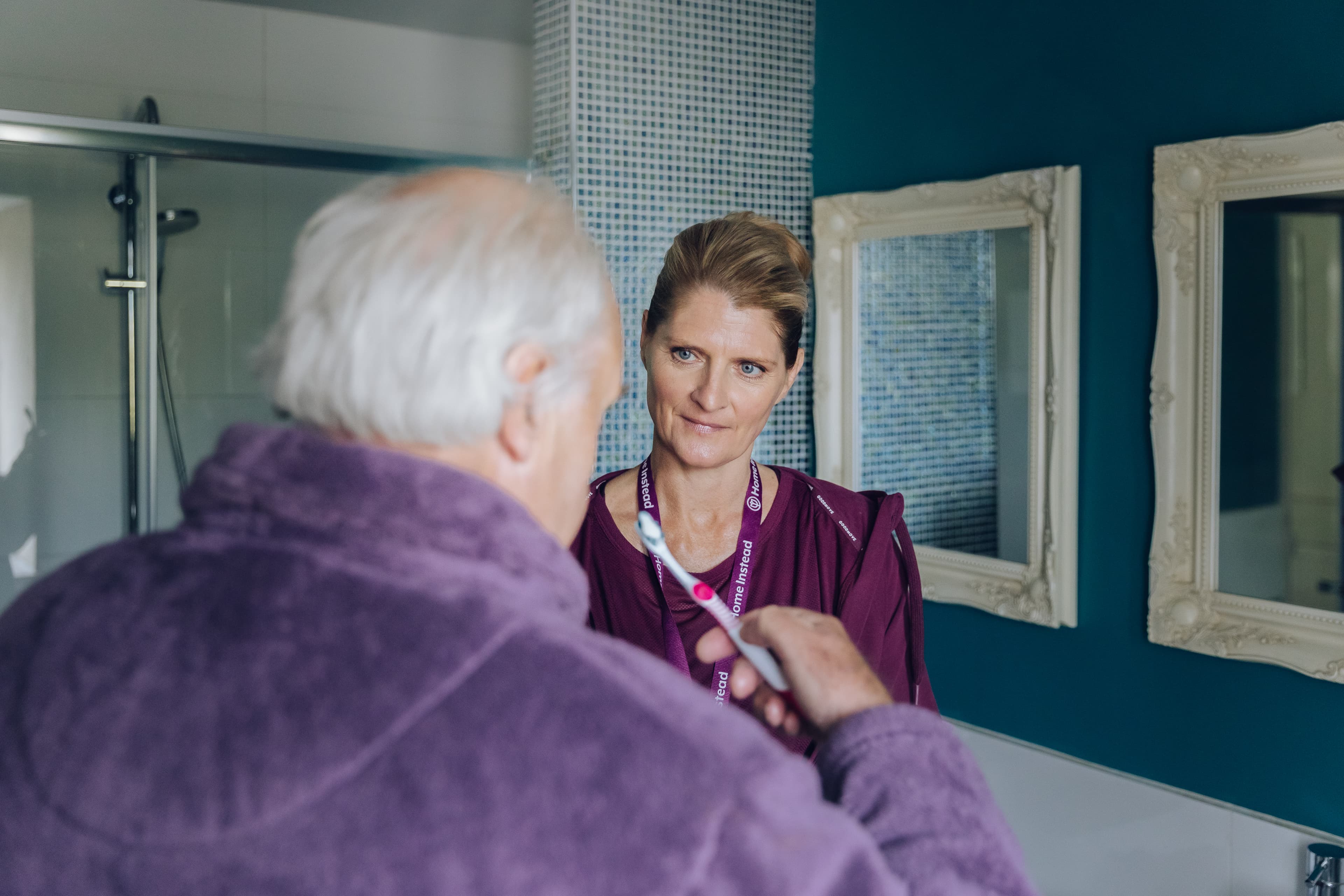 An older man with white hair brushes his teeth as a woman looks on, standing in a bathroom with blue walls and mirrors. - Home Instead