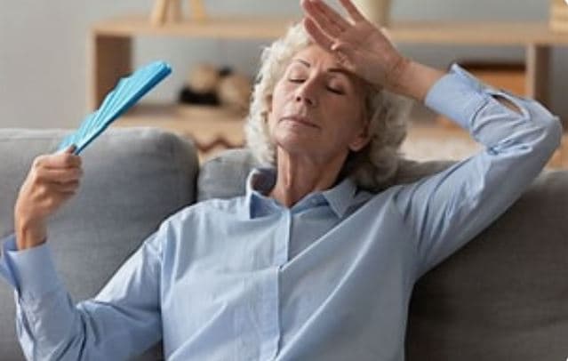 Elderly woman sitting on a couch, fanning herself with a blue fan, and appearing to feel hot or tired. - Home Instead
