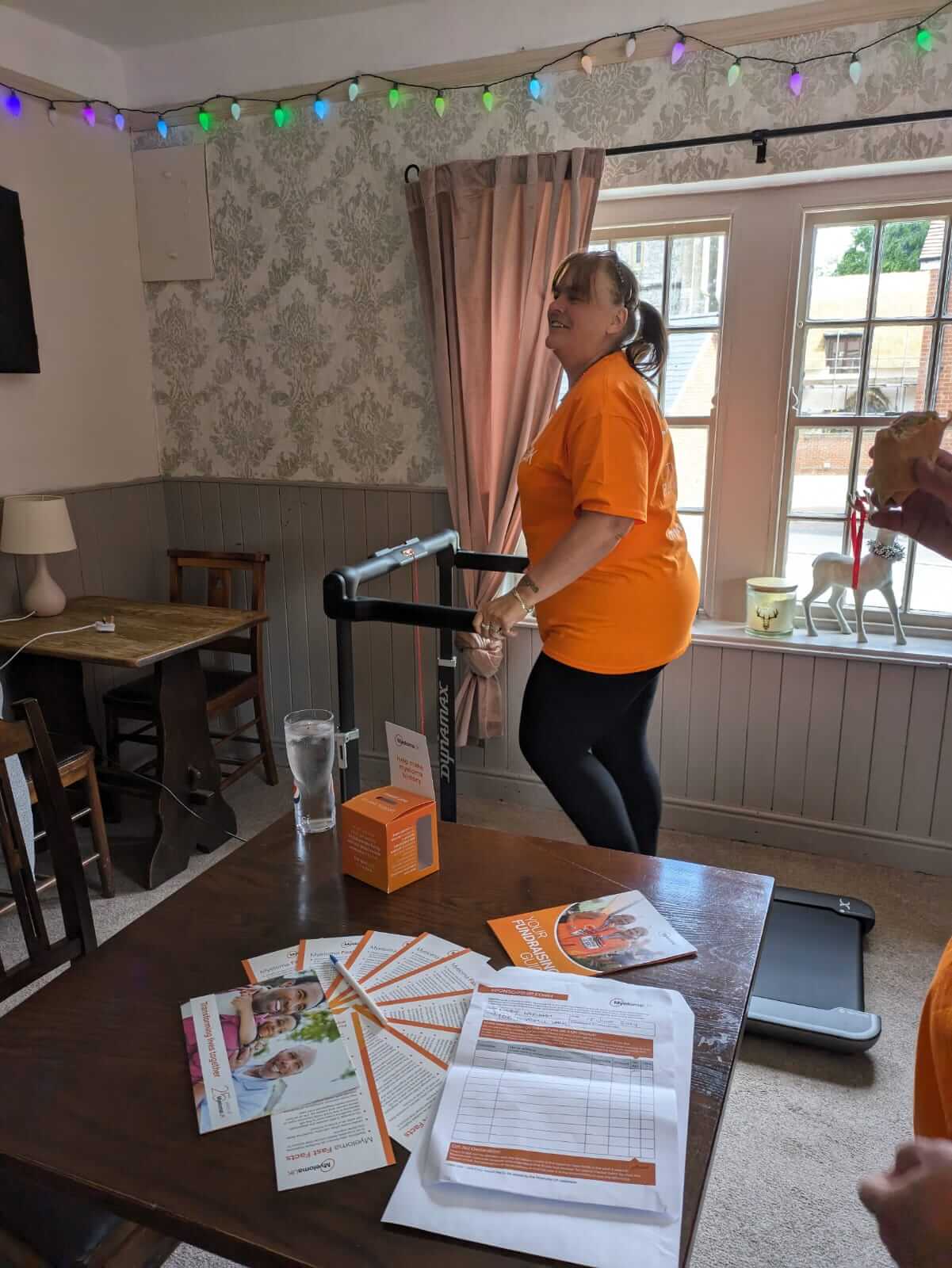 A woman in an orange shirt uses a treadmill desk in a cozy room with leaf-patterned wallpaper and colorful string lights. - Home Instead