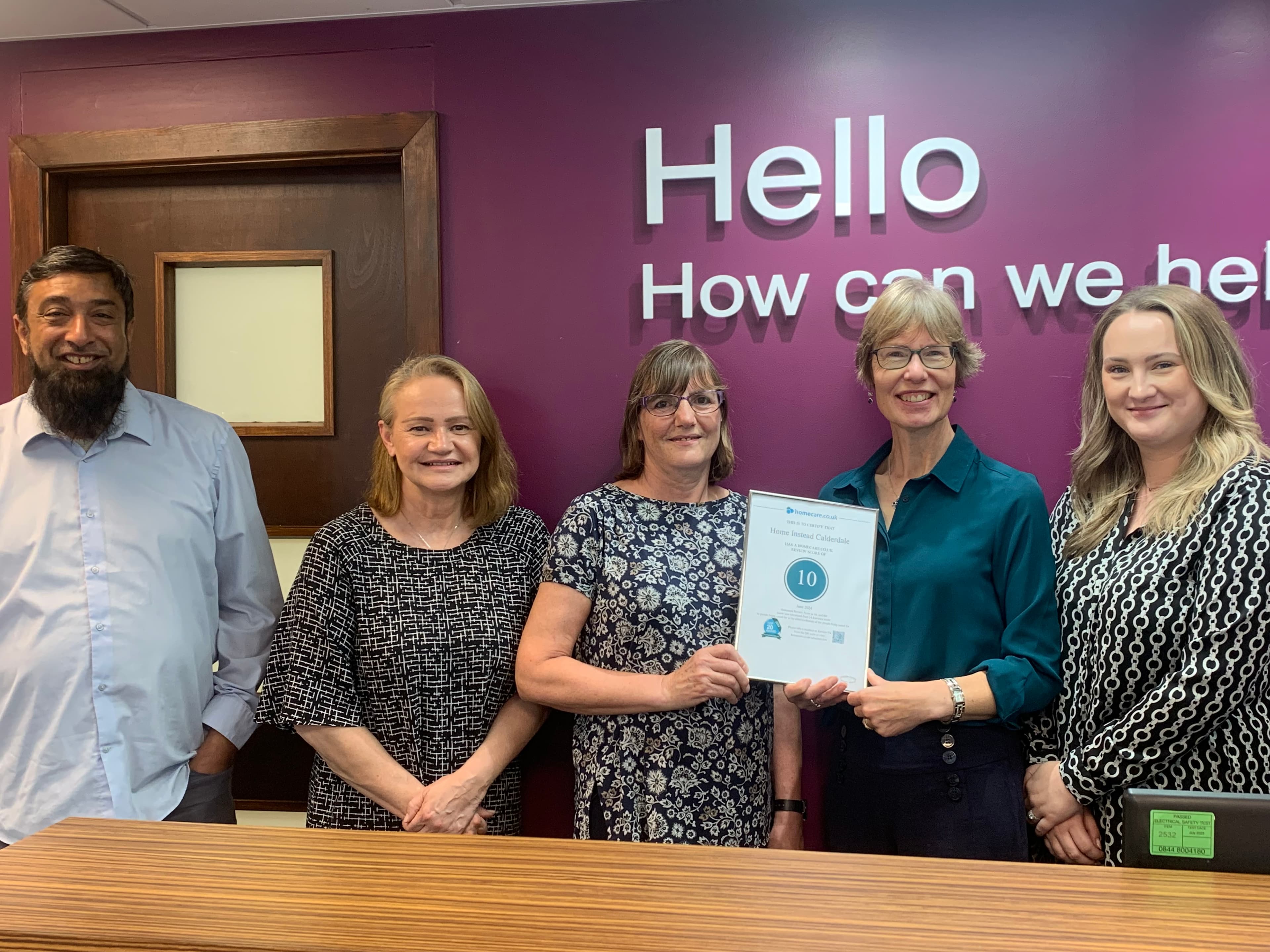 Five people standing together, one holding an award certificate, in front of a purple wall with the words "Hello, How can we help?. - Home Instead