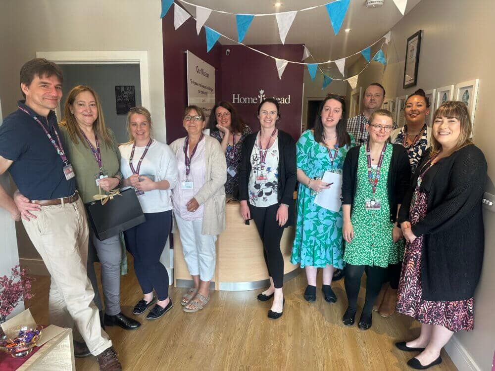 A group of people smiling and posing together in an office environment decorated with bunting. - Home Instead